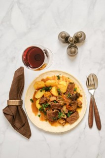 A plate holds a serving of beef stew with potatoes. A glass of red wine is nearby. A napkin is