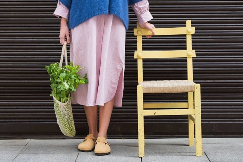 A stylish woman in a pink striped dress and blue sweater holds a mesh bag filled with fresh greens.