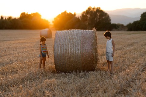 Two young children, one boy and one girl, play near a large hay bale in a field as the sun sets,