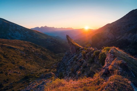 A stunning sunset casts warm hues over a rugged mountain landscape, highlighting rocky peaks and