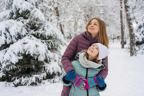 A loving mother embraces her smiling daughter in a winter wonderland. Both are dressed warmly,