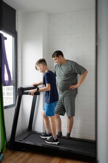 A parent supervises a child using a treadmill in a modern home gym. The room is bright with natural