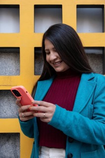 A Latin woman smiles while looking at her smartphone. She's in a blue coat and stands against a