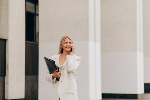 A confident businesswoman in a white suit chats on her smartphone while holding a folder outside a