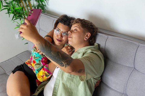 A lesbian couple relaxes on a gray sofa, capturing a selfie with a smartphone. They appear