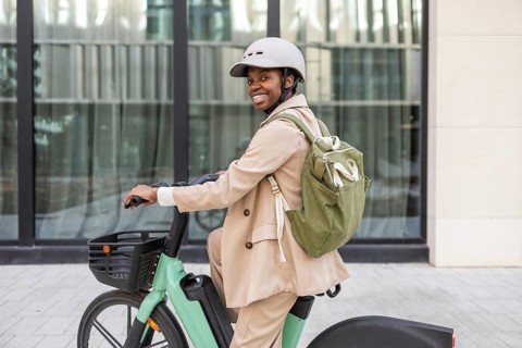 A black woman rides a bicycle in an urban environment, wearing a helmet and backpack She exudes