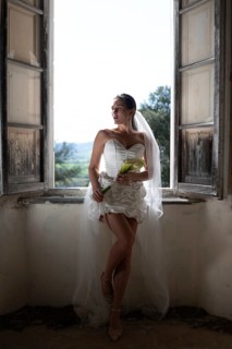 A bride in an elegant white dress and veil poses by a rustic window in an ancient villa in Treia,