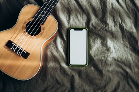 A smart phone with a blank screen and a ukulele rest on a crumpled bed sheet, illuminated by