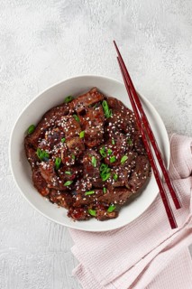 Crispy beef, with green onions and sesame seeds, in a bowl with chopsticks, on a textured surface,
