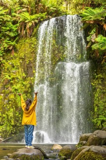 A woman in a yellow sweater stands with arms raised, admiring a waterfall surrounded by green