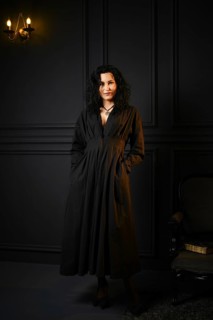 A woman stands in a dimly lit room. She wears a long black dress and has curly hair. Her hands are