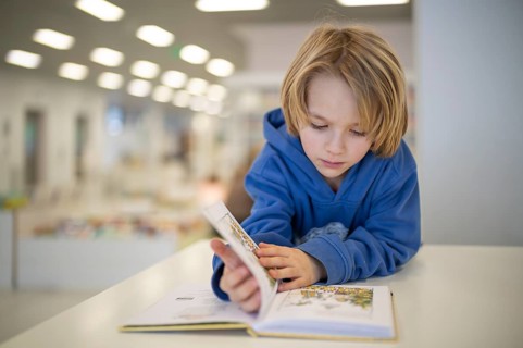 Interior shot, boy, 6 years old, blonde, reading book in the children's section of the city