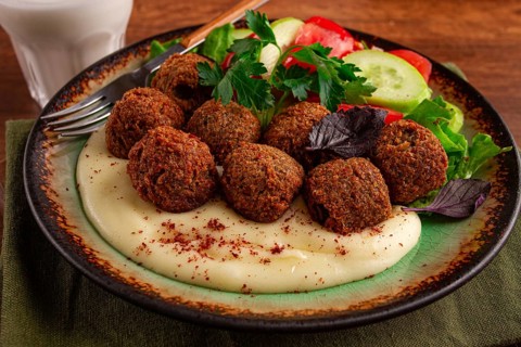 A plate holds falafel balls, tahini, salad with tomatoes and cucumbers, and a drink. The setting is