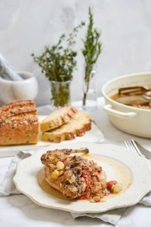 Rabbit stew is placed on a white plate next to slices of bread. There are herbs in small vases in