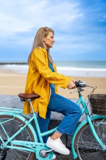 Young woman wearing yellow raincoat sitting on her light blue bicycle, enjoying the view of the sea