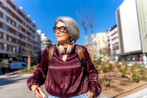Middle aged woman with chic gray hair and stylish eyeglasses, wearing headphones and a backpack,