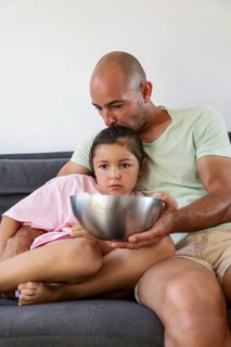 A father and his young daughter sitting on a couch at home, sharing a bowl of popcorn. The scene