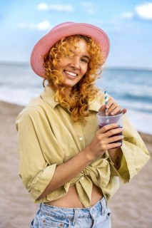 Young woman with curly red hair and pink hat smiling and enjoying a refreshing drink on a sunny