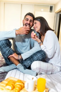 Young couple wearing pajamas, sitting in bed and eating donuts for breakfast, with orange juice and