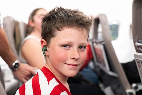 A young passenger with earbuds sits comfortably in an airplane seat, wearing a red and white