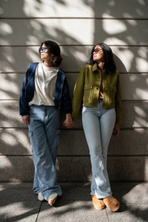 Two lesbian women stand confidently against a wall, holding hands. They wear stylish casual outfits