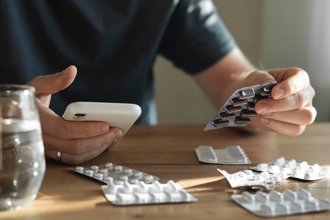 A man holds a package of pills in one hand and a smartphone in the other, sitting at a wooden table