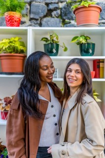 Two young businesswomen smiling together in front of a shelf adorned with vibrant plants,