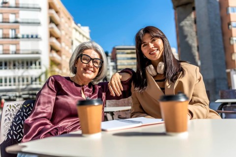 Two smiling women, an older mother and younger daughter, sitting at an outdoor cafe table with