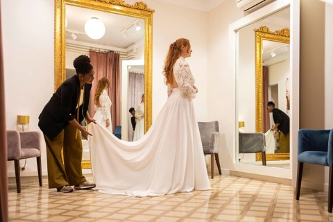 A bride tries on a wedding dress in a boutique, assisted by a stylist. The room features ornate