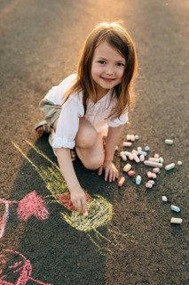 A young girl smiles while drawing on asphalt with colorful chalk. The sun casts warm light,