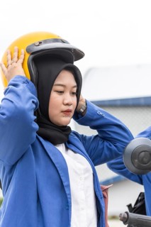 Asian Muslim woman wearing a hijab and a blue jacket, adjusting a yellow helmet, prepares to ride a