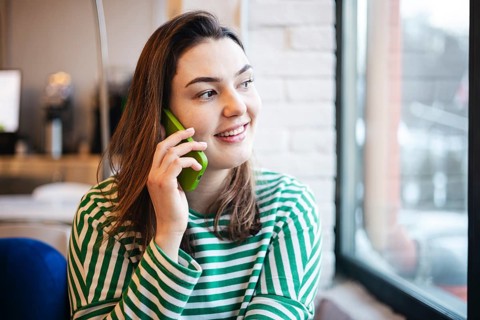 A cheerful woman enjoys a coffee break in a cafe while chatting on her smartphone, dressed in a