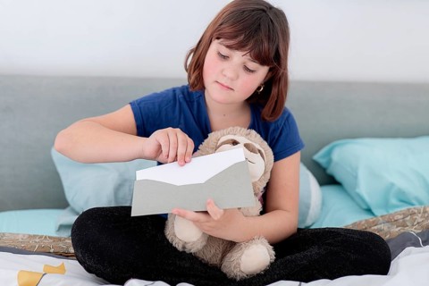 A young girl, holding a teddy bear and opening a letter on the bed, possibly containing a gift from