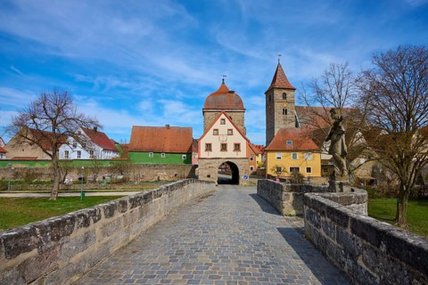 Historic stone bridge leads to a village with church and city gate under a clear blue sky, spring,