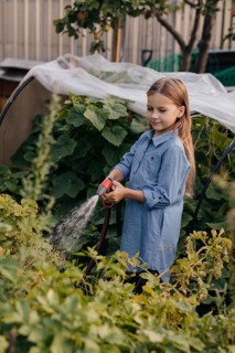 A young girl in a blue dress waters plants in a small greenhouse. Surrounded by greenery, she tends