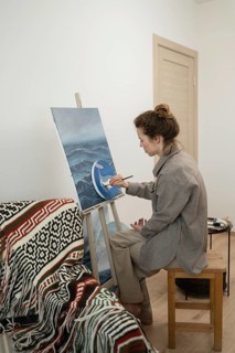 A woman sits on a wooden stool, painting on a canvas set on an easel. The room is cozy, with a