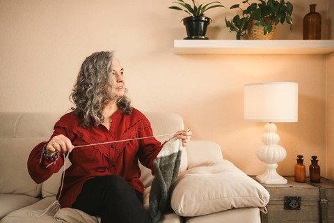 A woman in a red blouse knits on a beige sofa, enveloped in cozy decor. Houseplants and soft