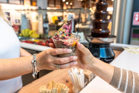 Vendor's hand serving a colorful frozen yogurt dessert with fruit and sprinkles in a plastic cup to