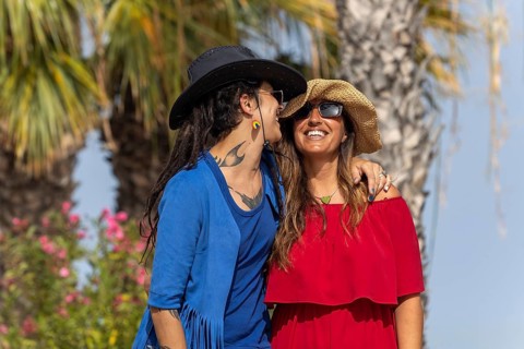 A joyful lesbian couple shares a moment of laughter under the sun, surrounded by palm trees and