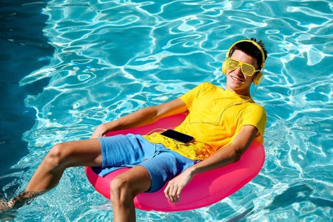 A young teenage boy relaxes on a pink pool float, wearing yellow sunglasses and headphones. He