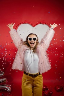 A joyful woman throws her arms in the air amidst falling confetti, celebrating Valentine's Day. She
