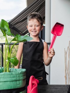 A cheerful young boy stands on a balcony, holding a red gardening shovel. Dressed in a black apron,