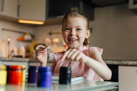 A happy child in a light pink top paints with vivid colors in a sunlit kitchen. The scene exudes