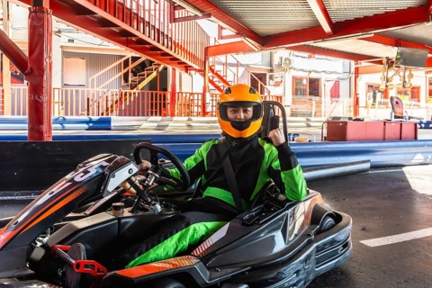 Man in orange helmet and green racing suit sits confidently in go kart on track, raising a fist in