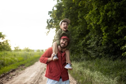 A joyful dad carrying his son on his shoulders strolls through a lush green field. The pair enjoys