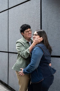 A joyful curvy lesbian couple shares a tender moment, embracing outdoors against a modern gray wall