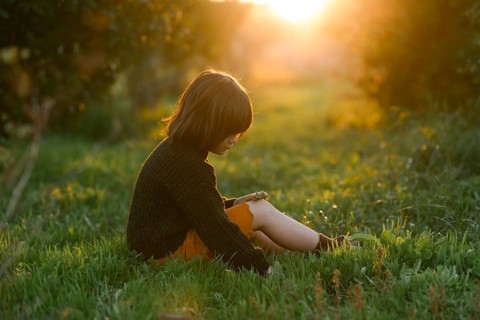 A child sits in a green field with orange trees, bathed in the golden glow of the setting sun. The