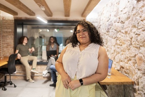 A confident female professional stands in the foreground of a modern office, while a diverse team