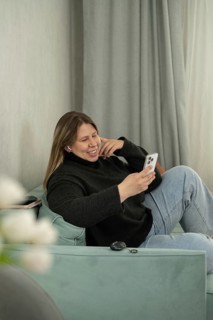 A cheerful woman sits comfortably on a pastel-colored sofa, using her smartphone and smiling.