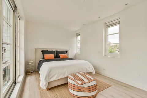 A bright, minimalist bedroom features a cozy bed with orange and black cushions, large windows